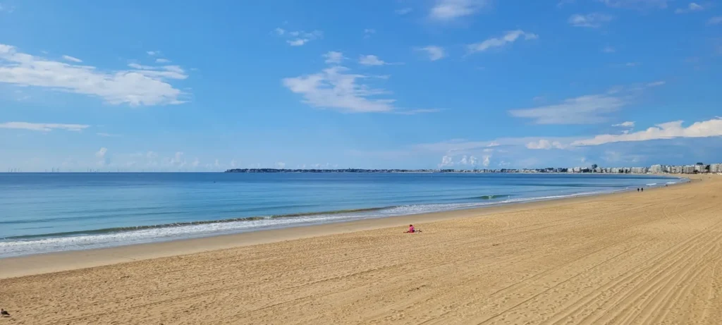 Grande plage de sable fin de La Baule sous un ciel bleu avec nuages. Vue panoramique sur l'océan Atlantique et la côte urbaine au loin, cadre idéal pour un séminaire d'entreprise avec l'Agence Helper.