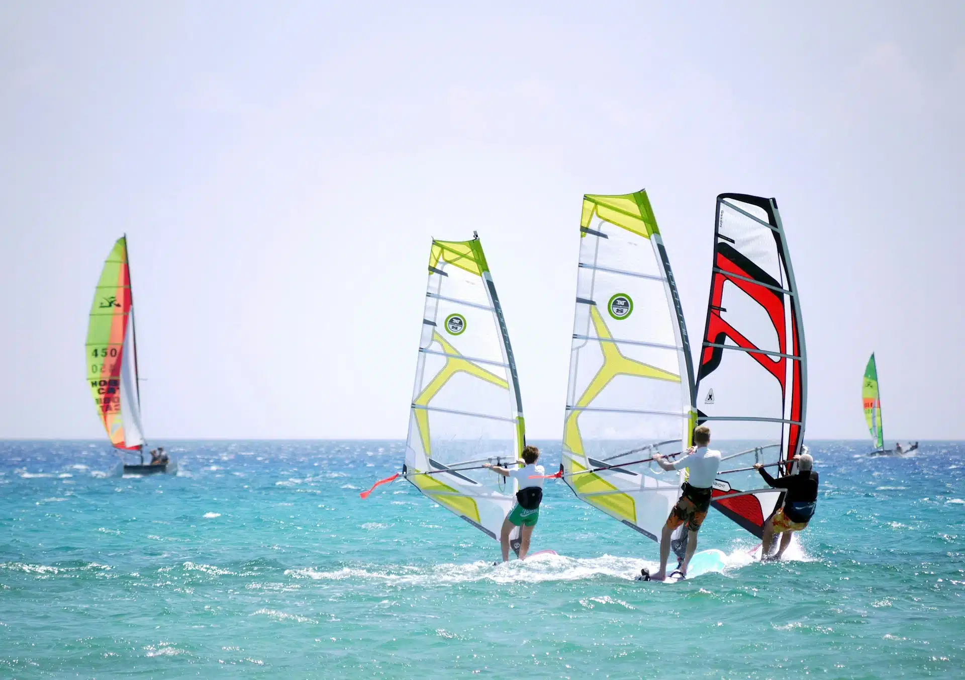 Trois planches à voile en action sur une mer turquoise, avec un voilier Hobie Cat en arrière-plan, illustrant une activité de Team Building à La Baule organisée par l'Agence Helper.