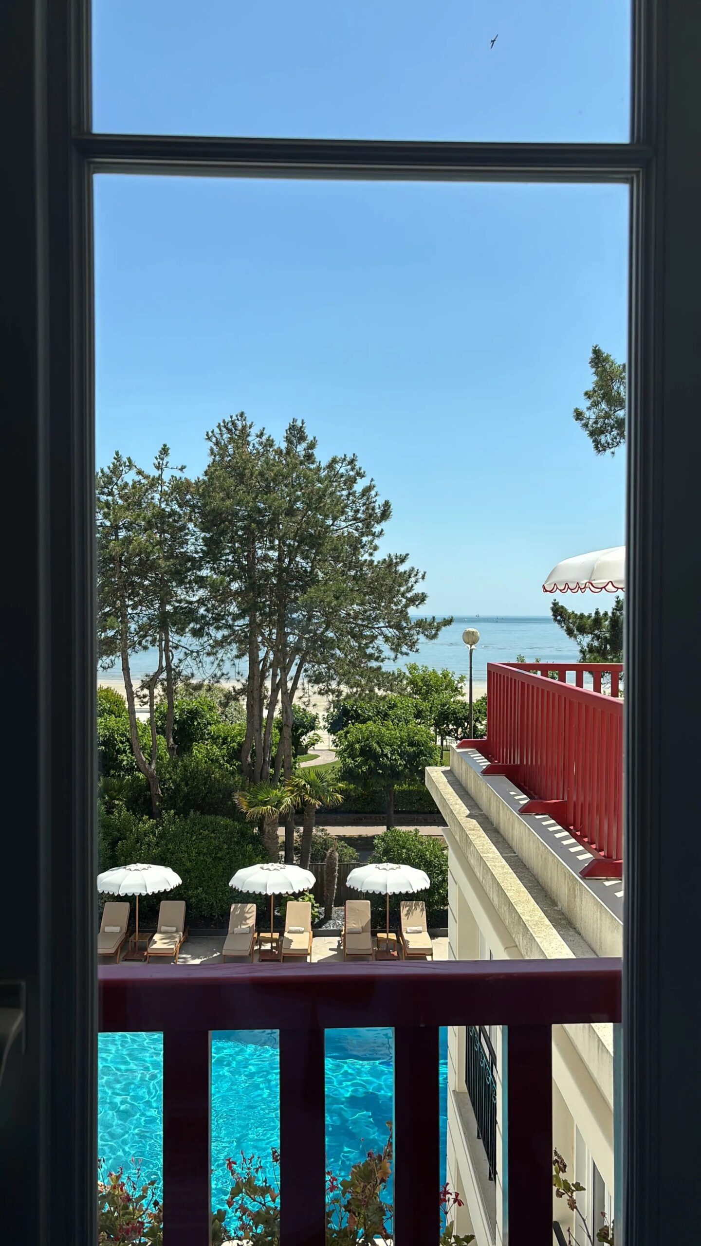 Vue depuis une fenêtre d'hôtel à La Baule sur une piscine azur, des parasols blancs et la plage de sable fin bordant l'océan Atlantique sous un ciel bleu.