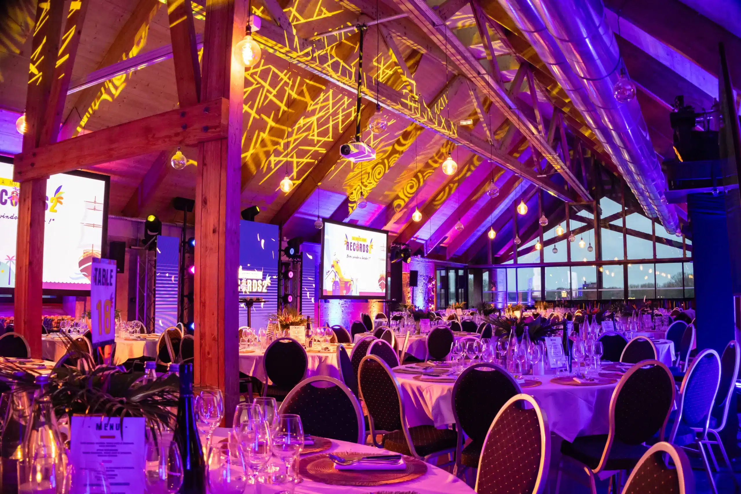 Salle d'événement professionnel à Nantes dressée par l'Agence Helper : tables rondes dressées, éclairage d'ambiance violet et écrans géants pour une soirée clé en main.